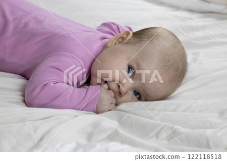 Cute and beautiful 4-month-old baby girl laying on bed and look at the camera. Infant in pink bodysuit on bed against white background. 122118518