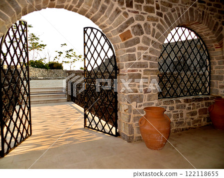 Stone entrance arch and windows with metal bars and gates. Red ceramic jug Stone entrance arch and windows with metal bars and gates. Red ceramic jug 122118655