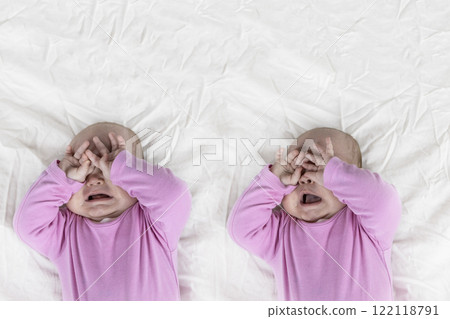 Set of two photos of 4-month-old baby girl laying on bed and crying. Infant  in pink clothes wants to sleep and rubs eyes 122118791