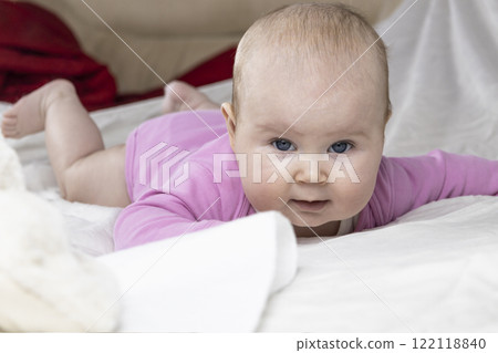 Cute and beautiful 4-month-old baby girl laying on bed and look at the camera. Infant in pink bodysuit on bed against white background. 122118840