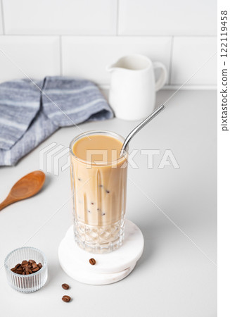 Cold latte coffee in a tall glass with milk and straw on a light table on a white brick background Cold latte coffee in a tall glass with milk and straw on a light table on a white brick background 122119458