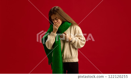 Young girl in beige sweater and green scarf, suffering from inflammation, cold and flu symptoms, coughing isolated on red background. 122120255