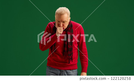 Senior man in red sweater and scarf coughing, suffering from cold and flu symptoms on green studio background. Throat irritation 122120258