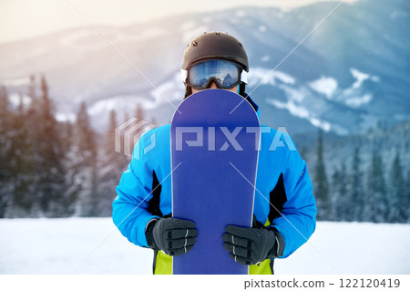 Portrait Of Man On The Background Of Mountains And Blue Sky, Holding Snowboard And Wearing Ski Glasses. Ski Goggles Of Snowboarder With The Reflection Of Snowed Mountains. Winter Sports Portrait Of Man On The Background Of Mountains And Blue Sky, Holding Snowboard And Wearing Ski Glasses. Ski Goggles Of Snowboarder With The Reflection Of Snowed Mountains. Winter Sports 122120419