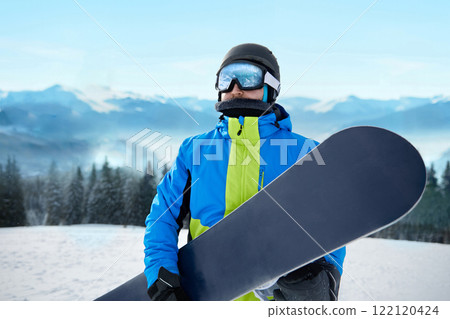 Snowboarder Of Man At Ski Resort On The Background Blue Sky, Hold Snowboard. Wearing Ski Glasses. Ski Goggles With The Reflection Of Snowed Mountains 122120424