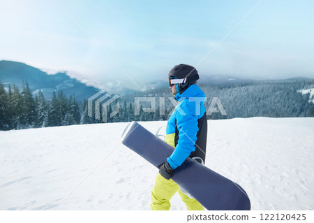 Snowboarder Of Man At Ski Resort On The Background Blue Sky, Hold Snowboard. Wearing Ski Glasses. Ski Goggles With The Reflection Of Snowed Mountains. Snowboarder Of Man At Ski Resort On The Background Blue Sky, Hold Snowboard. Wearing Ski Glasses. Ski Goggles With The Reflection Of Snowed Mountains. 122120425