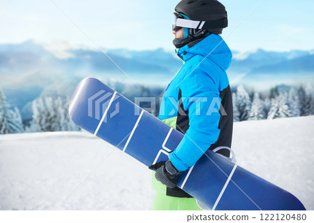 Snowboarder Of Man At Ski Resort On The Background Blue Sky, Hold Snowboard. Wearing Ski Glasses. Ski Goggles With The Reflection Of Snowed Mountains Snowboarder Of Man At Ski Resort On The Background Blue Sky, Hold Snowboard. Wearing Ski Glasses. Ski Goggles With The Reflection Of Snowed Mountains 122120480