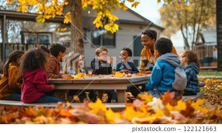 Happy diverse children and teacher sitting around a table outdoors in autumn, surrounded by fallen leaves. 122121663