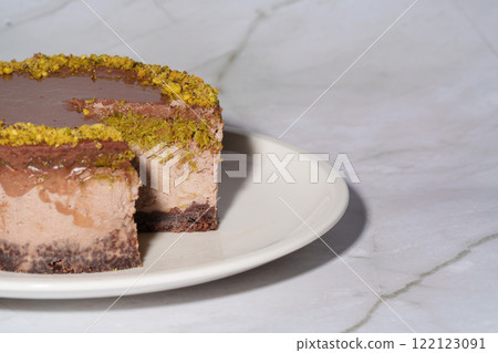 Close up Dubai chocolate cheesecake with pistachio paste and kataifi covered in chocolate ganache on white plate. White marble background with copy space. Indulgence, gourmet desserts, art of culinary 122123091