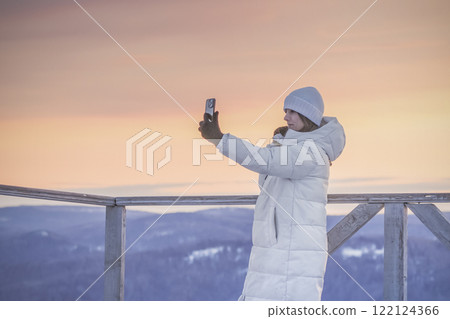 A woman n a white down jacket takes a selfie against the sunset in the mountains, A woman joyfully taking a selfie in a stunning snowy landscape during a breathtaking sunset 122124366