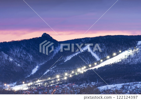 Evening panorama mountain ski resort. illuminated ski slope with burning lights during a beautiful lilac sunset. Dramatic and Beautiful Night Ski Resort Landscape Filled with Twinkling Lights in the 122124397