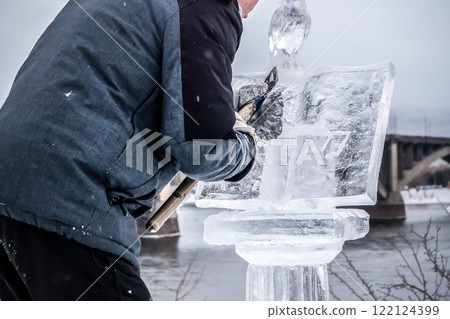 A man carves an icy transparent figure out of ice with a cutter, a neat and subtle professional creative work outdoors in winter. A skilled winter ice sculptor meticulously works on a stunning piece A man carves an icy transparent figure out of ice with a cutter, a neat and subtle professional creative work outdoors in winter. A skilled winter ice sculptor meticulously works on a stunning piece 122124399