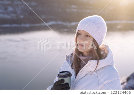 A woman in a white jacket and hat drinks hot coffee in winter by the riverbank and enjoys the moment and silence. A Winter Woman Happily Enjoying a Warm Beverage While Relaxing Near a Beautiful Frozen A woman in a white jacket and hat drinks hot coffee in winter by the riverbank and enjoys the moment and silence. A Winter Woman Happily Enjoying a Warm Beverage While Relaxing Near a Beautiful Frozen 122124406