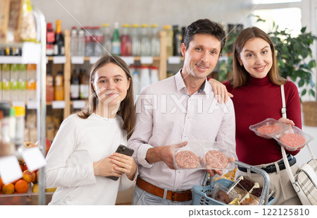 Dad, mom and daughter choose raw cooled semi-finished hamburger cutlet in shop 122125810