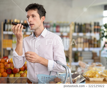 European man with basket of groceries in hands stands in store and dictates audio message 122125822