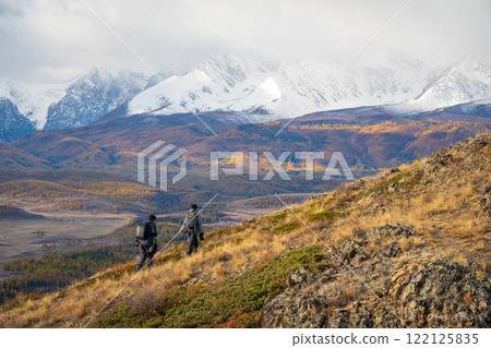 Two hikers explore a scenic mountain valley with snowy peaks and autumn colors 122125835