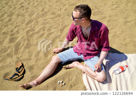 A Russian young man in a plaid shirt and shorts sitting on the sandy beach in summer, wearing sunglasses. Nearby are a bottle of water, flip-flops, and seashells A Russian young man in a plaid shirt and shorts sitting on the sandy beach in summer, wearing sunglasses. Nearby are a bottle of water, flip-flops, and seashells 122126694