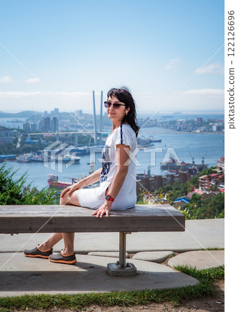A 50-year-old Russian woman in a white dress sits on a bench in Nagorny Park, Vladivostok, looking at the camera with Golden Horn Bay in the background 122126696