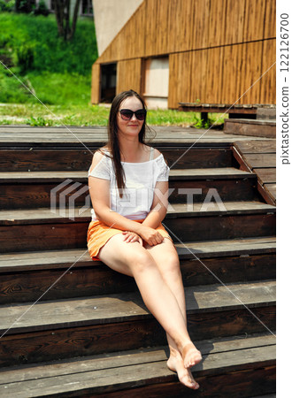 A 35-year-old Russian woman sits barefoot on wooden stairs in summer, wearing sunglasses, an orange skirt, and a white top, smiling slightly outdoors 122126700