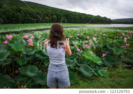 A young Russian woman photographs a lotus lake on her phone, focusing on mobile photography and blogging for social media content outdoors in nature 122126703