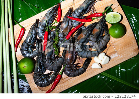 Tiger shrimp are carefully arranged on a wooden board, surrounded by vibrant red chilies, green limes, and aromatic herbs. This culinary display captures the essence of Asian flavors. 122128174