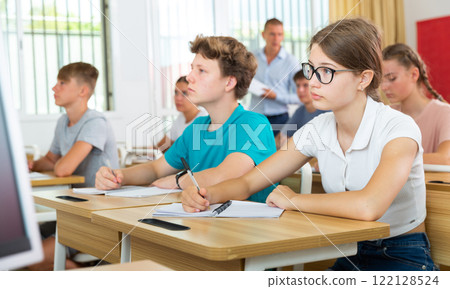 Teen girl in glasses listening to lecturer and writing in notebook in classroom 122128524