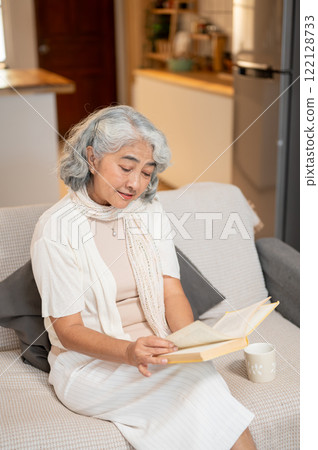 A relaxed Asian retired senior woman enjoys reading a book on a sofa in the living room. 122128733