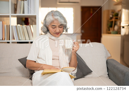 A well-being, happy Asian senior retired woman reads a book and enjoys a hot drink on a sofa. 122128734
