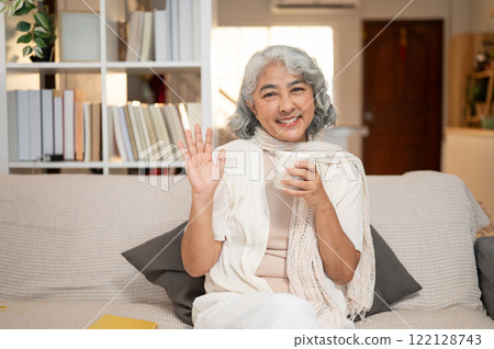 A beautiful Asian senior woman sits on a sofa with a coffee mug in her hand, waving at the camera. 122128743