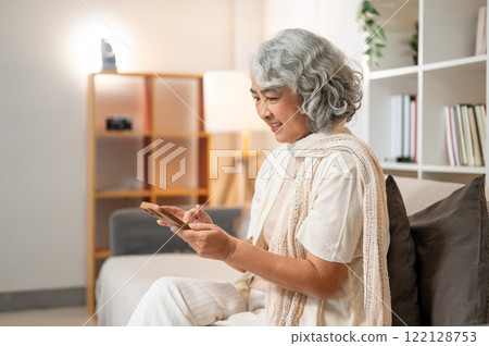 A happy Asian senior woman uses her smartphone to chat on social media while sitting on a sofa. 122128753