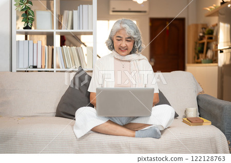 A happy Asian senior woman uses a laptop while sitting on a sofa in the living room. A happy Asian senior woman uses a laptop while sitting on a sofa in the living room. 122128763