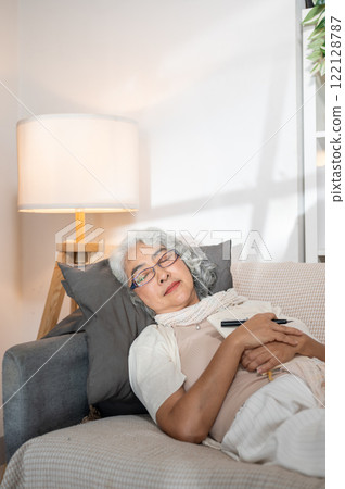 A relaxed Asian senior woman falls asleep while writing her diary on the sofa at nighttime. A relaxed Asian senior woman falls asleep while writing her diary on the sofa at nighttime. 122128787