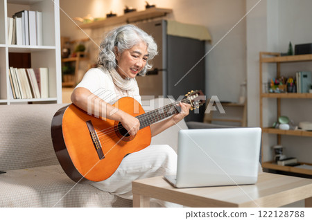 A relaxed Asian senior woman practices a guitar in the living room while studying an online lesson. A relaxed Asian senior woman practices a guitar in the living room while studying an online lesson. 122128788