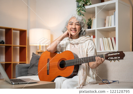 A beautiful, happy Asian retired woman sits on a sofa in the living room with her acoustic guitar. 122128795