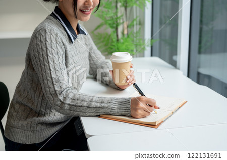 A close-up of a smiling Asian woman writes her diary and enjoys a hot drink in a cafe. 122131691