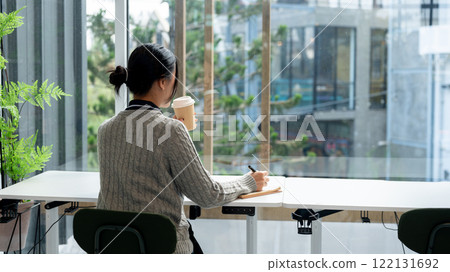 A back view of a relaxed and calm Asian woman sitting at a table by a window in a cafe. 122131692