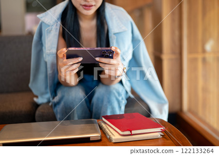 A close-up of a woman in a denim jacket sits in a coffee shop, using her smartphone, watching videos 122133264