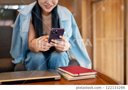 A smiling Asian woman sitting in a coffee shop, enjoying using social media on her smartphone. 122133266