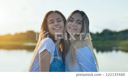 Teenage friends hugging, laughing near lakeside during golden sunset light, sharing warm friendship moment 122135480
