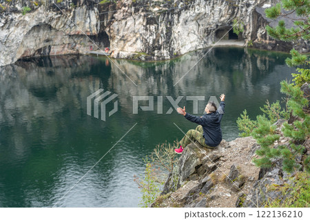 Young man sitting on a rock with raised arms, enjoying the view of a flooded marble quarry Young man sitting on a rock with raised arms, enjoying the view of a flooded marble quarry 122136210
