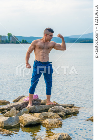 Strong sportsman flexing biceps while standing on rocks at lake in summer, enjoying nature and healthy lifestyle Strong sportsman flexing biceps while standing on rocks at lake in summer, enjoying nature and healthy lifestyle 122136216