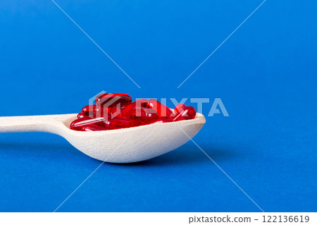 Vitamin capsules in a spoon on a colored background. Pills served as a healthy meal. Red soft gel vitamin supplement capsules on spoon 122136619