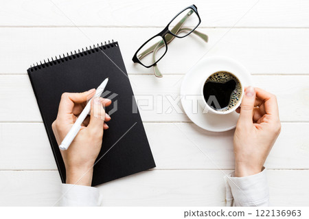 Woman hand with pencil writing on notebook and hold coffee cup. Woman working on office table with coffee 122136963
