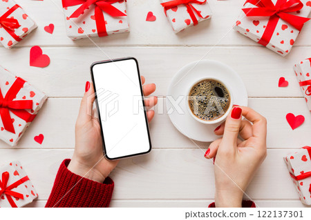 Female hands holding smart phone with coffee for Valentine day, gift box and envelope, hearts 122137301
