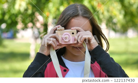 Teenage girl taking pictures with pink and beige wooden toy camera in a park, playing and having fun 122137430