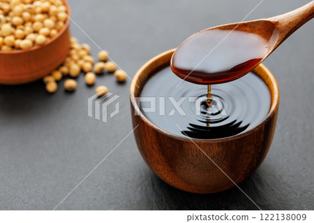 soy sauce pouring from spoon in bowl with splash and drop and dry soybeans on black stone background 122138009