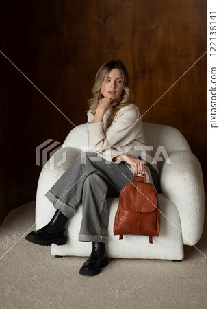 beautiful girl in gray pants and beige sweater posing with a small shiny brown leather backpack 122138141