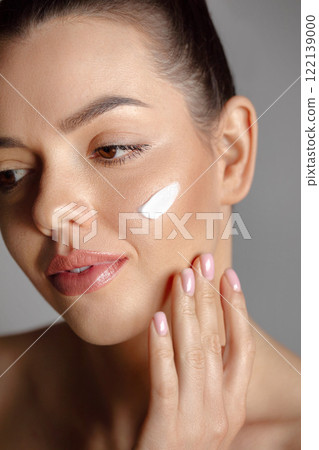 Beautiful woman applying white cream with fingers on cheek. Portrait of beauty model with natural nude make up and touching her face. Spa, skincare and wellness. Close up, copyspace. 122139000
