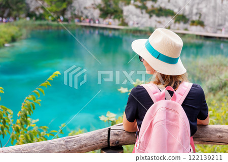 Back view young adult woman female traveler wearing pink backpack straw hat enjoy looking scenic view of turquoise waters Plitvice Lakes National Park, Croatia. Tranquil waterfall landscape trip tour 122139211
