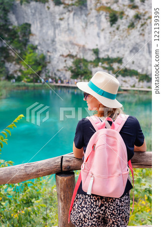 Back view young adult woman female traveler wearing pink backpack straw hat enjoy looking scenic view of turquoise waters Plitvice Lakes National Park, Croatia. Tranquil waterfall landscape trip tour 122139395
